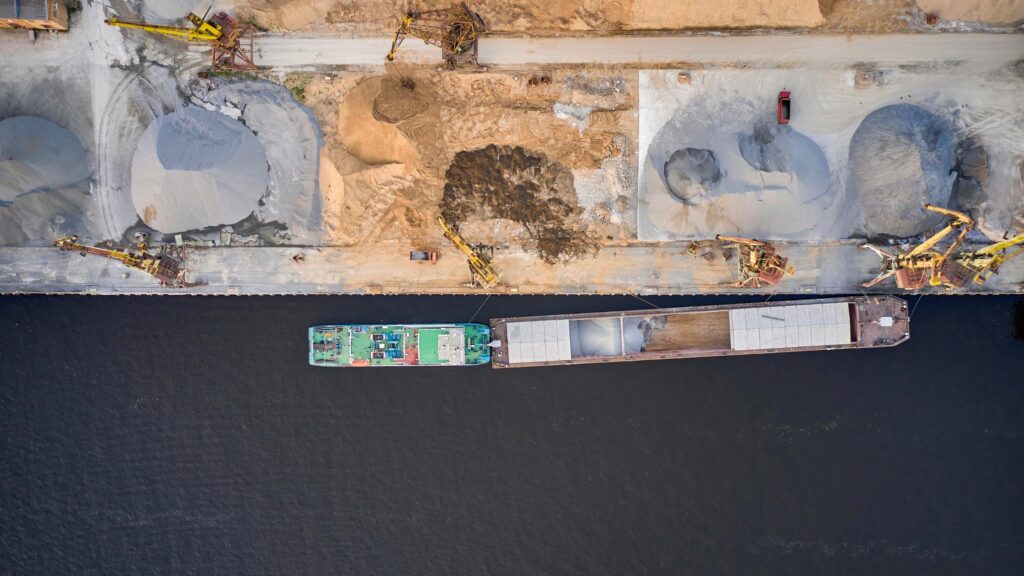 Aerial view of an industrial area along a waterfront. A large cargo ship is docked next to a concrete pier. Various construction machinery is present around the site of industrial material heaps next to dirt and concrete surfaces.