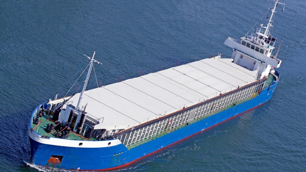 A large cargo ship with a blue hull and a white superstructure sails in the open water. The ship has a covered deck area for transporting goods. The image offers a clear overhead view of the vessel.