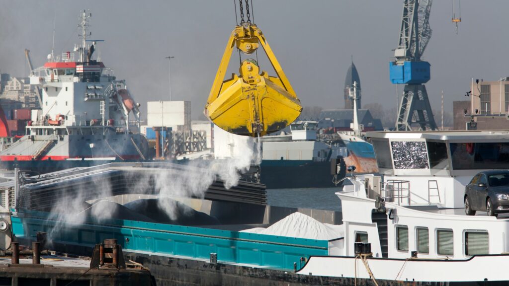 A cargo ship docked at a port is being loaded with powdered material using a yellow crane. Buildings, industrial structures, and other vessels are visible in the background. A car is parked on the deck of a nearby ship.