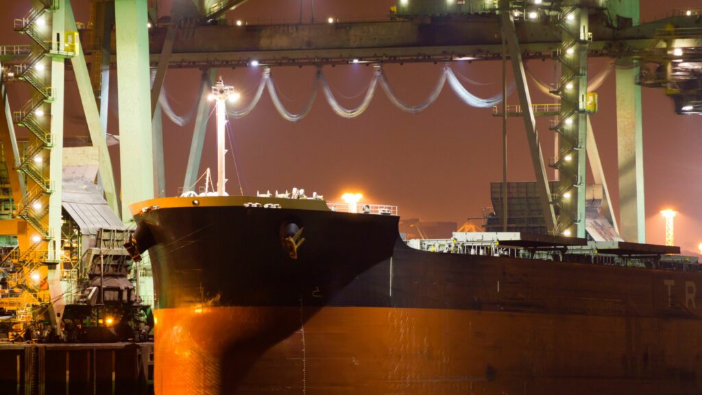 A large cargo ship is docked at a busy, well-lit port at night. The ship's bow is prominent in the foreground, while industrial cranes and equipment surround it. The scene is illuminated by various light sources, creating a vibrant atmosphere.