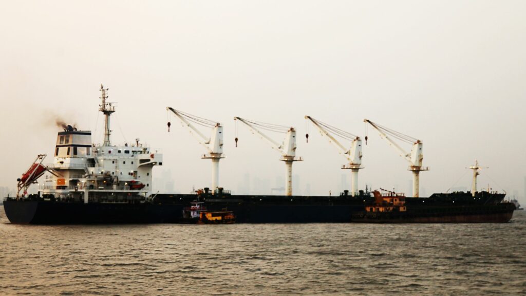 A large cargo ship with four cranes is anchored in calm waters. Smaller vessels are positioned alongside the ship. The sky is overcast, and the city skyline is faintly visible in the background.