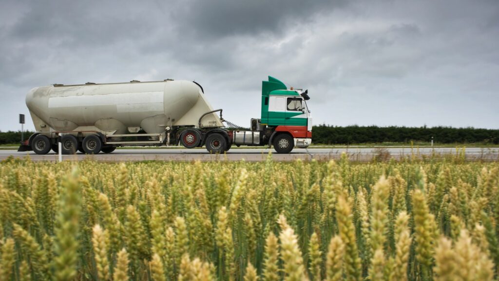 A green and white tanker truck drives along a highway under a cloudy sky. In the foreground, a field of wheat is visible. Trees line the horizon in the background.