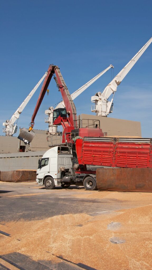 A red truck is parked near a large ship with cranes, as grain is being transferred from the ship into the truck. The scene takes place under a clear blue sky at a port or industrial area.