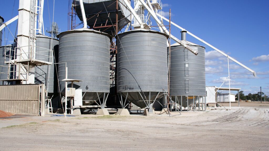 A row of large, gray metal grain silos stands on a dusty ground under a clear blue sky. Several pipes and ladders are attached to the silos, and a smaller building is visible in the background.