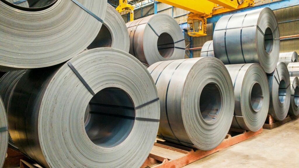 Large rolls of steel coils are stacked on wooden pallets inside an industrial warehouse. A yellow overhead crane is visible in the background, and metal beams support the structure's roof. The space is well-lit, highlighting the steel's gray surface.