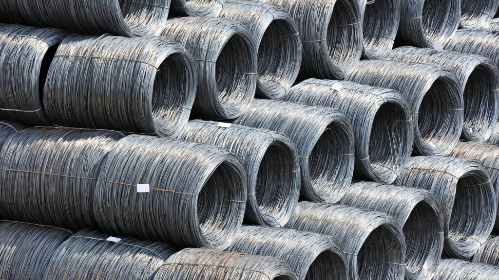 Stacks of industrial coiled steel wires are arranged in rows. The steel wires are tightly wound in large circular bundles, with each coil neatly positioned side by side, forming a uniform pattern across the image.