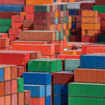 Stacks of colorful shipping containers are piled in a large outdoor storage area, reflecting global shipping routes' dynamic nature. The containers, primarily red, orange, green, and blue, form varied geometric patterns as they adapt to the post-pandemic world.