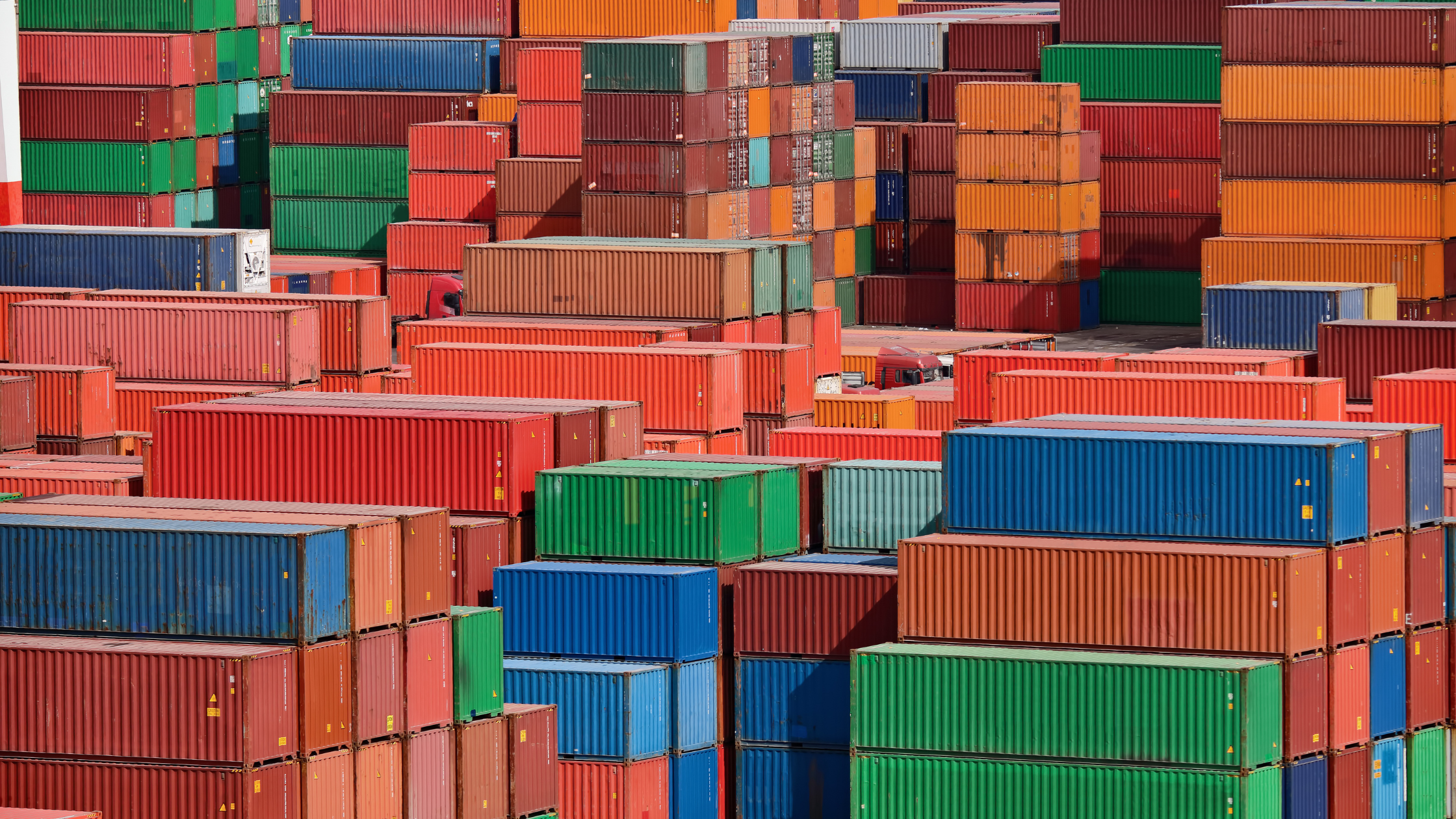 Stacks of colorful shipping containers are piled in a large outdoor storage area, reflecting global shipping routes' dynamic nature. The containers, primarily red, orange, green, and blue, form varied geometric patterns as they adapt to the post-pandemic world.