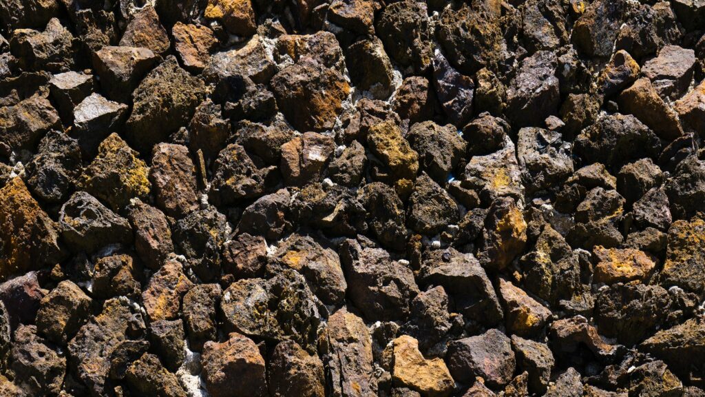 A close-up view of a collection of dark, rough textured rocks packed closely together. The surface features various shades of brown, gray, and black with a rugged appearance.