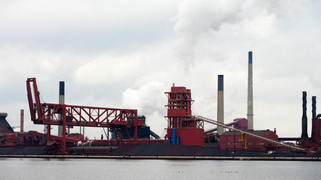 Industrial landscape with a steel mill by the water. Red structures, including a large conveyor system, dominate the scene. Several chimneys emit smoke into a cloudy sky, indicating active industrial processes. The foreground features a body of water.