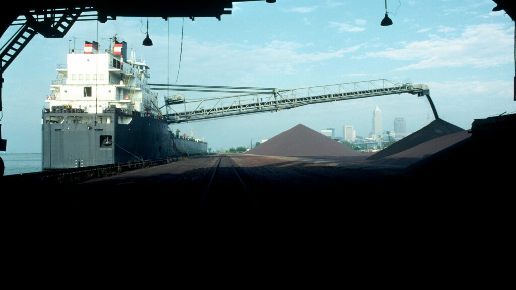 A large cargo ship is docked at a port, unloading a pile of dark granular material via an extended chute. The city skyline is visible in the background under a partly cloudy sky.