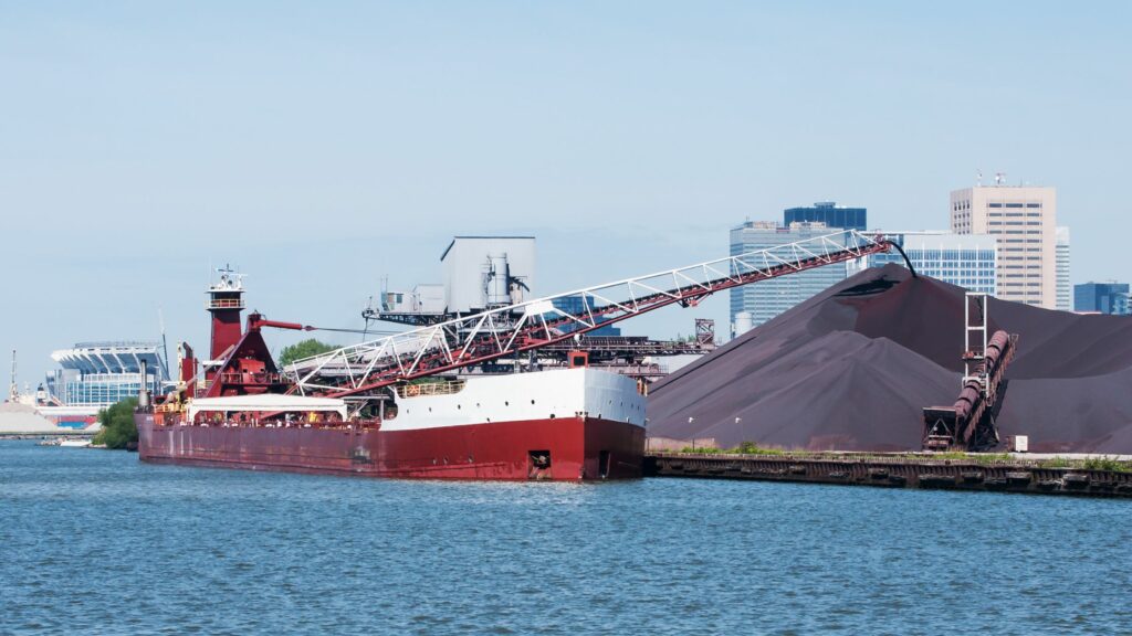 A large red cargo ship is docked at an industrial port. It is unloading dark-colored bulk material onto large piles via a conveyor system. The background features city buildings under a clear blue sky.