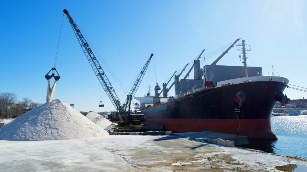 A large cargo ship is docked at a port. Two cranes are transferring piles of a light-colored bulk material, such as salt or sand, from the ship onto the dock. The scene is set on a clear, sunny day with a blue sky.