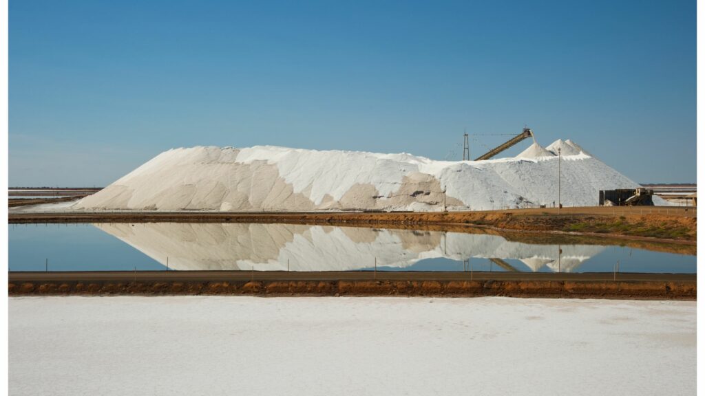 A large pile of harvested salt is reflected in a still body of water, with a conveyor belt on top of the mound. The sky is clear and blue, and the surrounding area is flat and industrial looking.