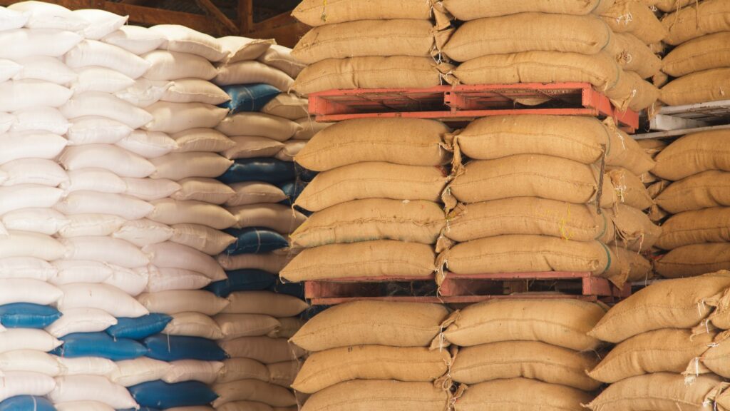 Stacks of burlap sacks, filled and organized on red pallets, are neatly arranged alongside white sacks with blue bases in a storage area. The sacks appear to be filled with bulk materials.