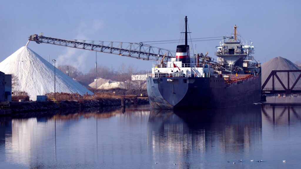 A large cargo ship is docked at an industrial port. A conveyor belt extends over a pile of white material, likely salt or minerals. Smoke rises from distant chimneys, and the sky is clear. The water reflects the scene, including seagulls nearby.